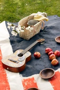 a guitar, peaches, and a picnic basket with dogwood flowers on a painted american flag picnic blanket.