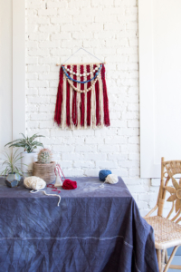 A red, cream, and blue woven flag tapestry hangs on a white brick wall above a blue tablecloth. There are a few balls of yarn and some houseplants on the table and a wicker chair is pulled out next to it.