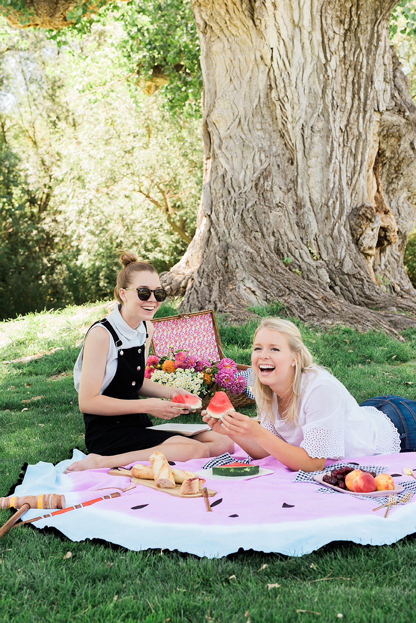 Two women sitting on a watermelon picnic blanket with snacks and flowers. They're in a shady park and they're laughing.