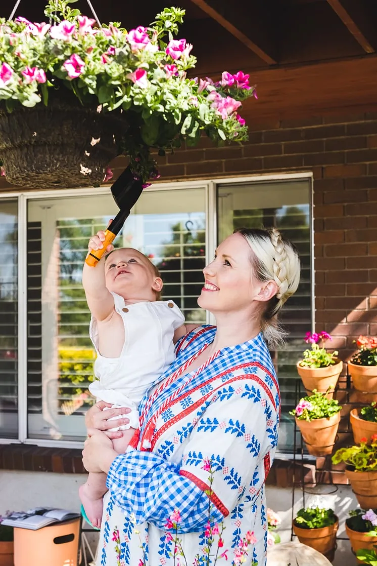 Brittany holds baby Jasper up to a hanging flower pot full of pink flowers. 
