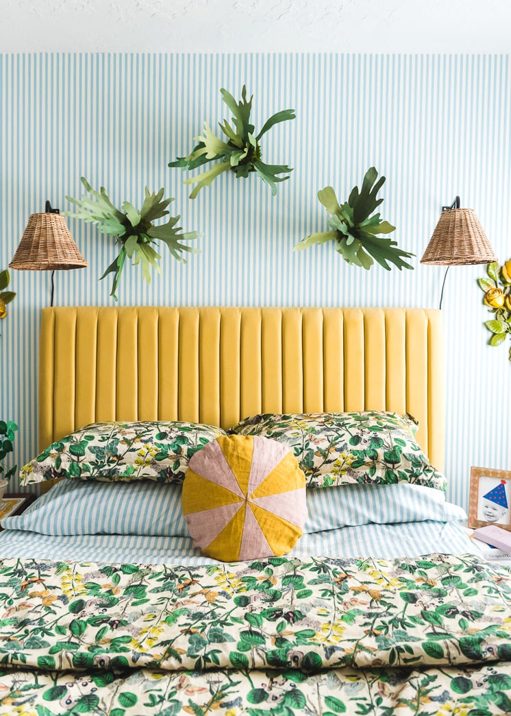 Interior shot of a bedroom with blue and white striped wallpaper. There's a yellow headboard, green floral bedding, paper staghorn ferns, and wicker lamps.
