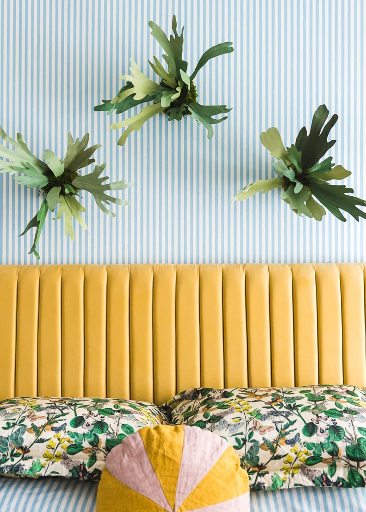 Interior shot of a bedroom with blue and white striped wallpaper. There's a yellow headboard, green floral bedding, paper staghorn ferns, and wicker lamps.
