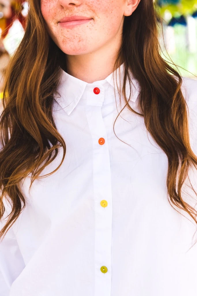 a white woman with brown hair wears a white blouse with rainbow buttons