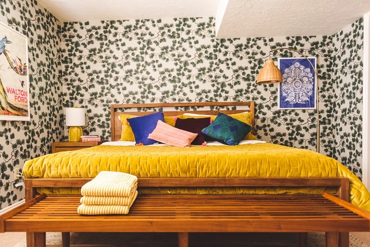 a room with pine-themed wallpaper. There are pillows in dark blue, pink, green, and black, the bed is warm wood with a mustard duvet, and there's a wicker lamp in the corner. There's also a blue art print on the wall.