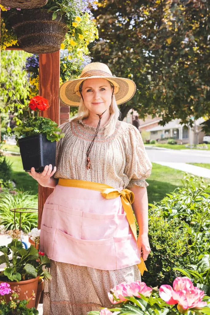 Brittany stands in the garden wearing an apron and a sun hat and holding a geranium.