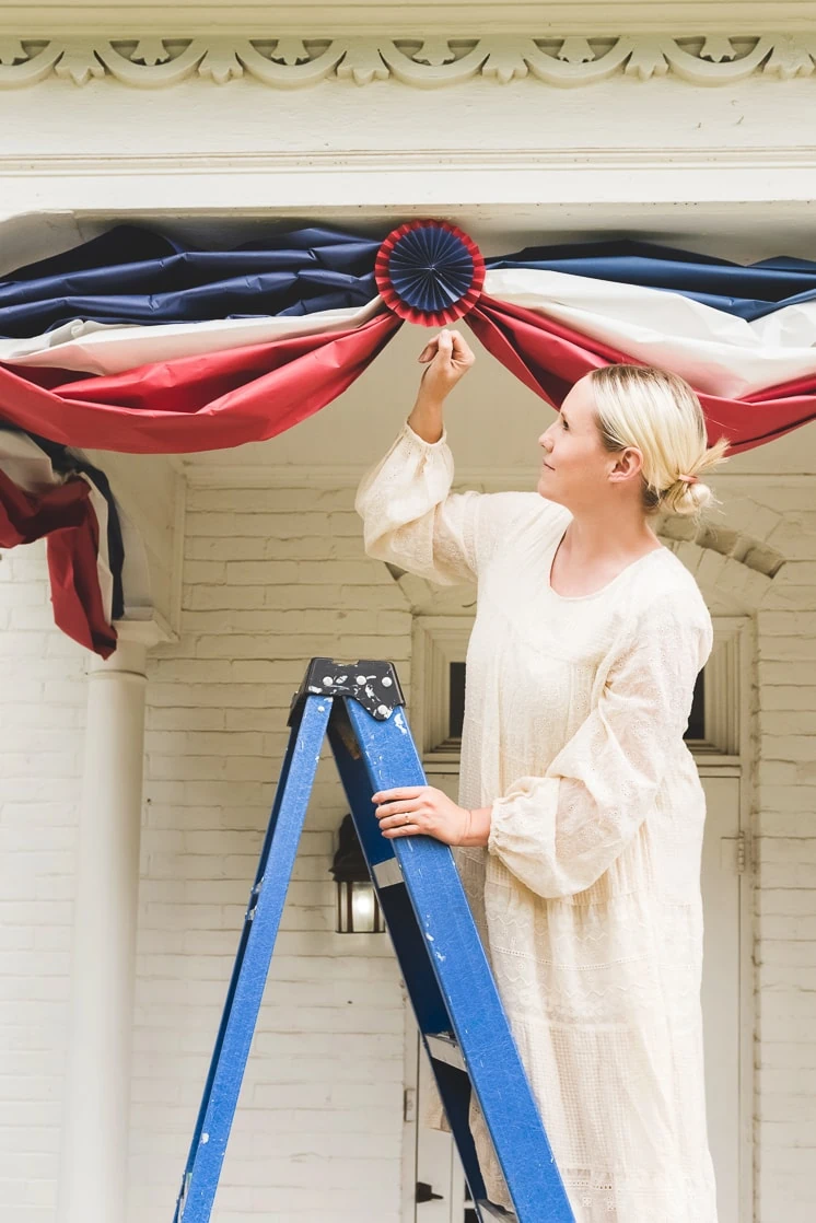 Brittany stands on a ladder and puts up red, white, and blue paper bunting on a white house. 
