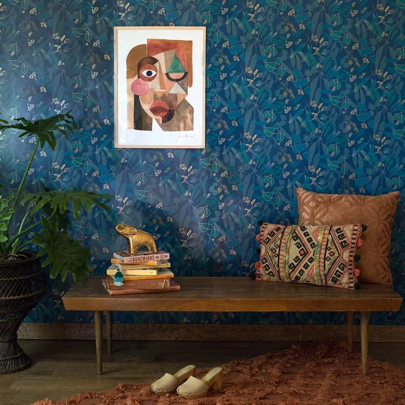 An interior photo. The wall is blue floral wallpaper, there's a big houseplant, a patterned rug, and pillows propped on a wooden bench.