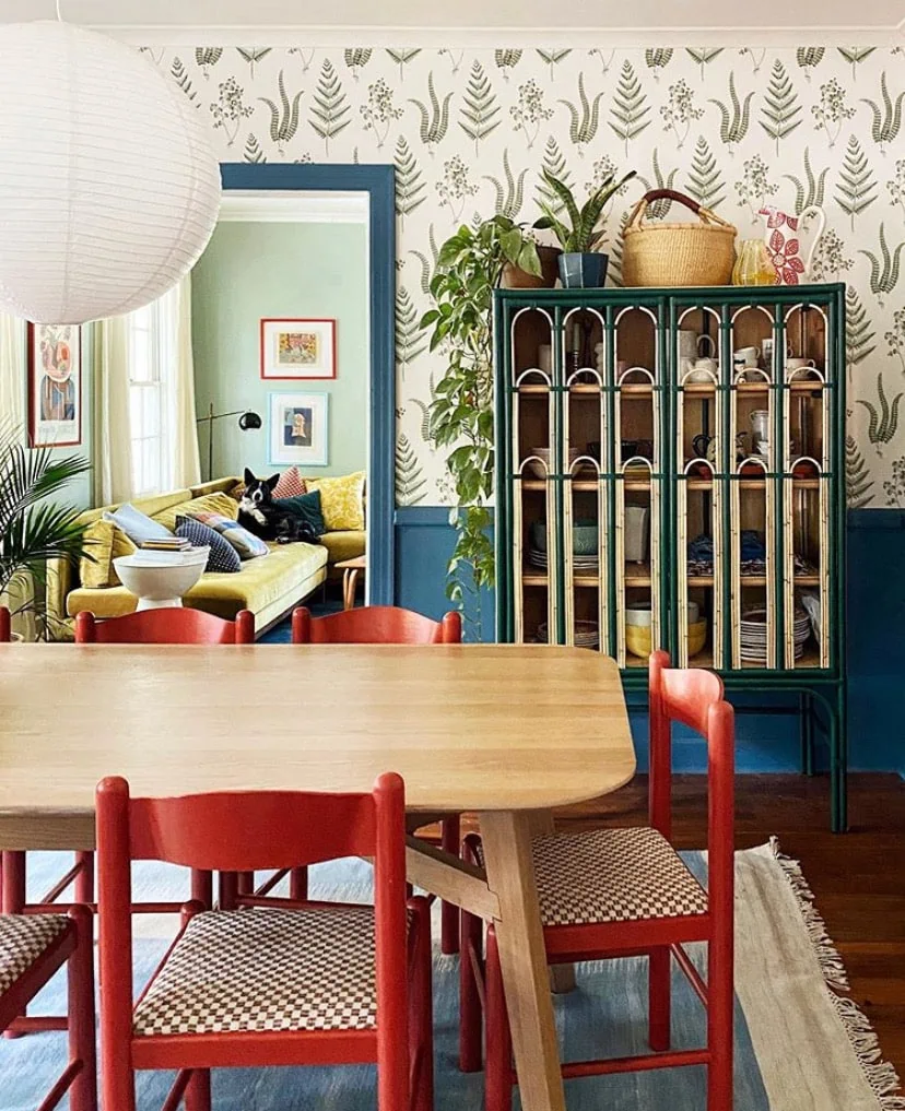 Interior shot of a colorful, eclectic dining room with red chairs, wallpaper and blue wainscoting, a green cabinet, and plants. 