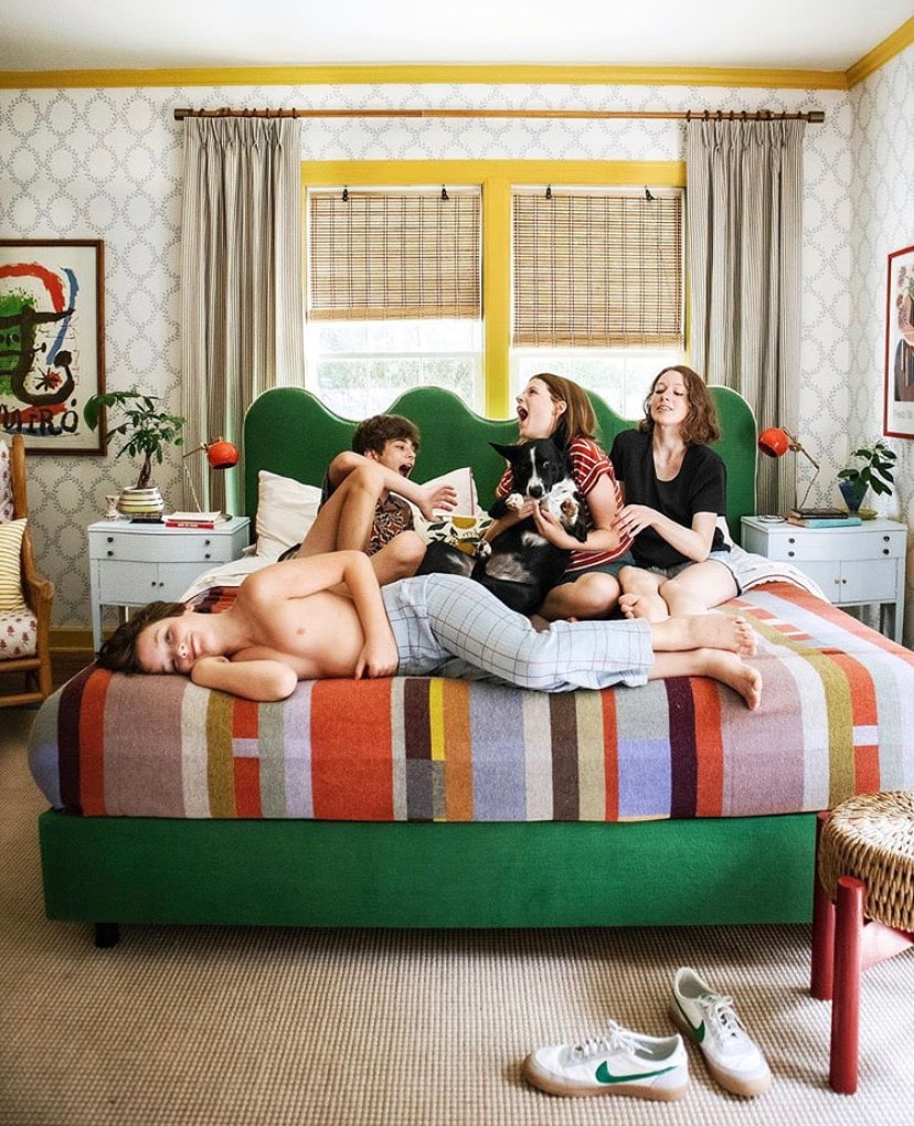 Four children lounging and laughing on a green bed with a striped quilt. One is holding a dog and there's a window behind them. 