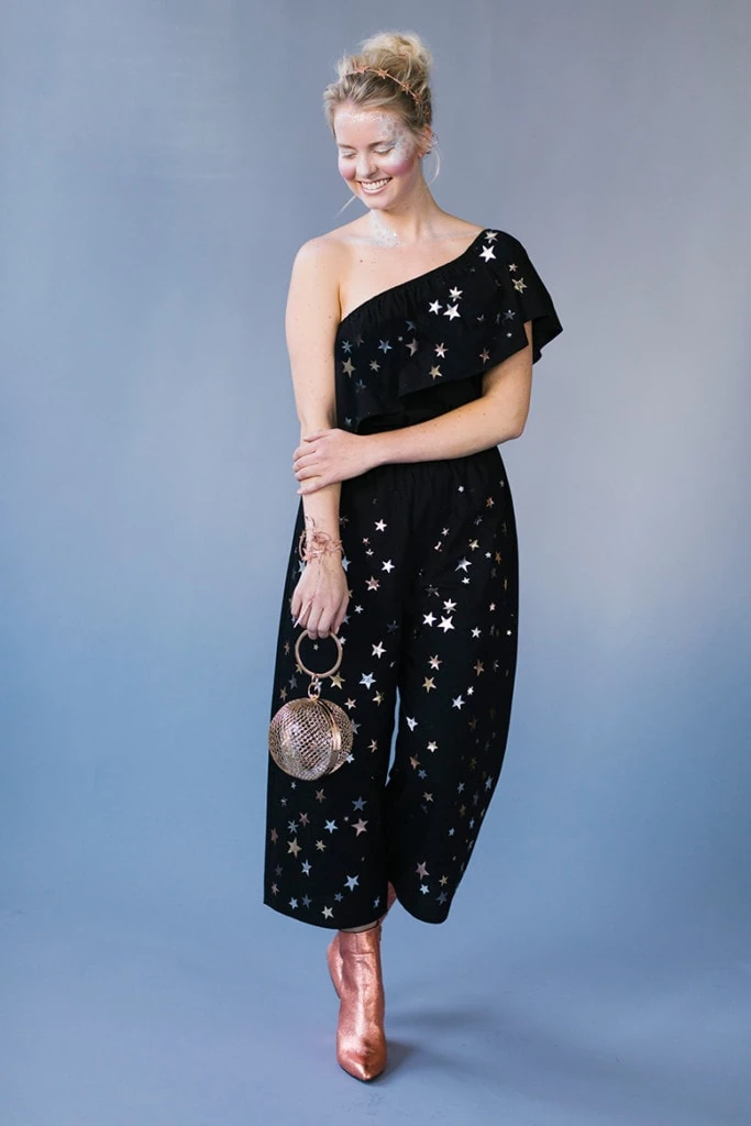 a blonde woman wearing an off-the-shoulder black jumpsuit with silvers stars all over it, sparkly makeup, and a spangled headband against a periwinkle background