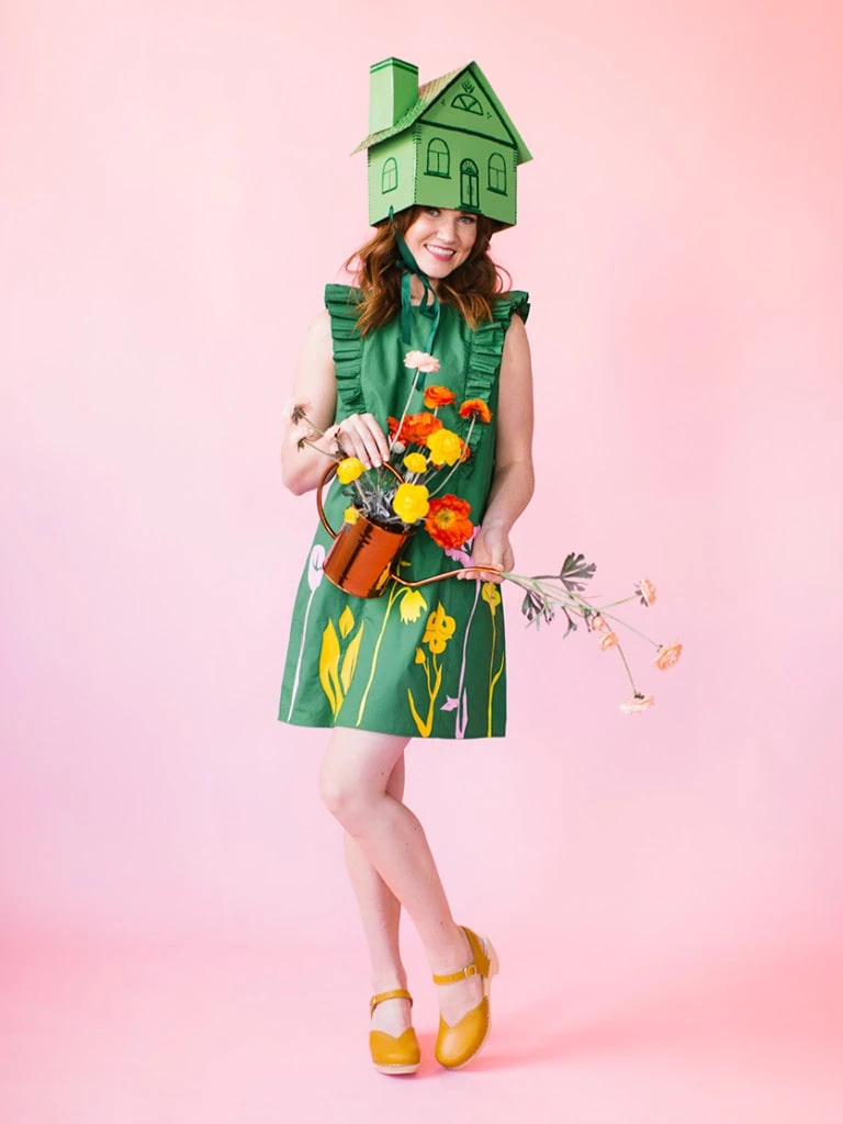a white woman with brown hair wears a green dress with iron on flowers, a greenhouse hat, and yellow clogs. She's holding flowers and a watering can.