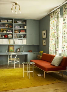 A library designed by Meta Coleman. It has soft sage green walls, a wooden floor, and a red couch.