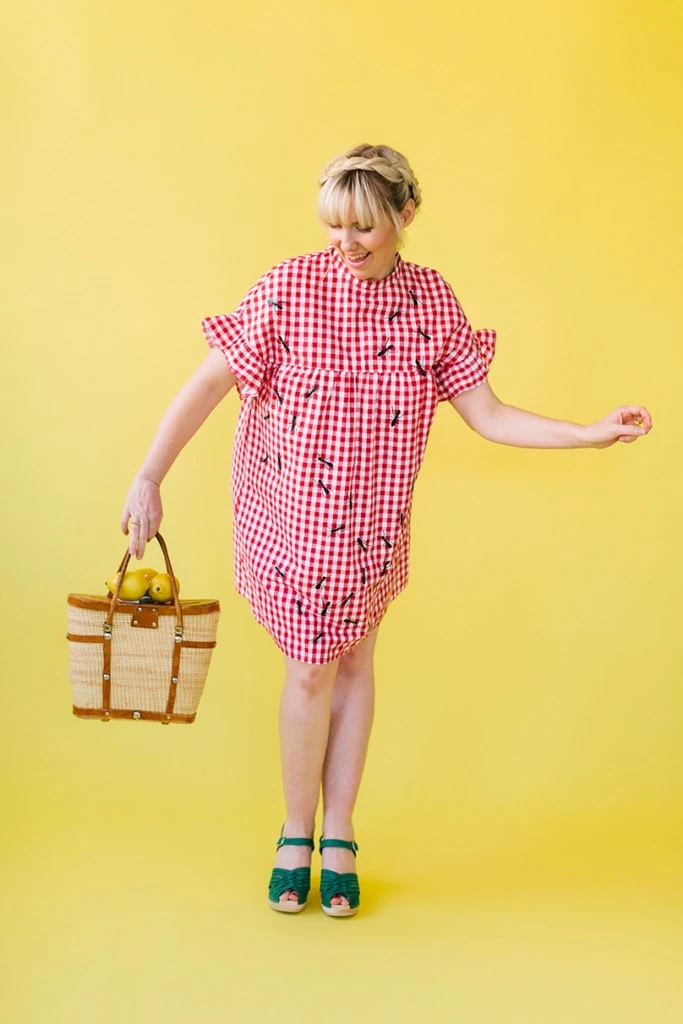 Brittany wears a gingham red dress with ants ironed onto it and holds a rattan picnic bag. 