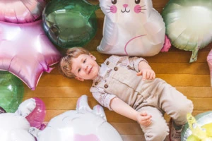 A little boy smiles up at the camera from a yellow rug, where he is lying. He's surrounded by Easter-themed balloons.