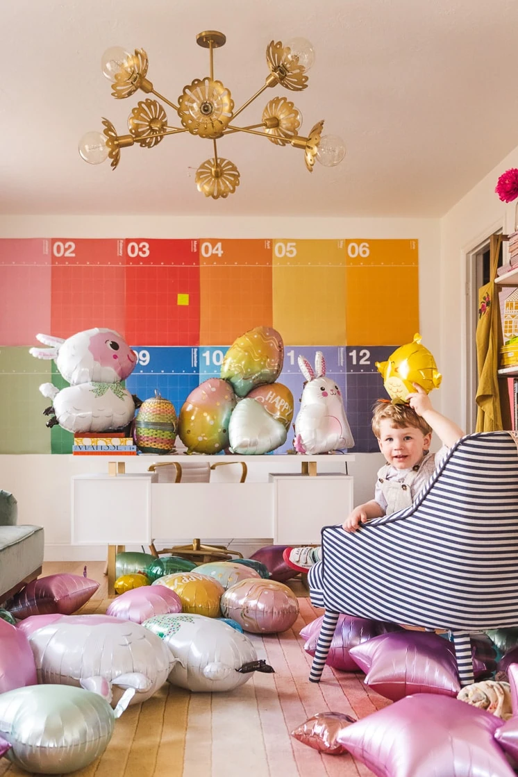 A little boy holds up a chick balloon. He's sitting on a chair in a colorful room strewn with Easter-themed balloons.