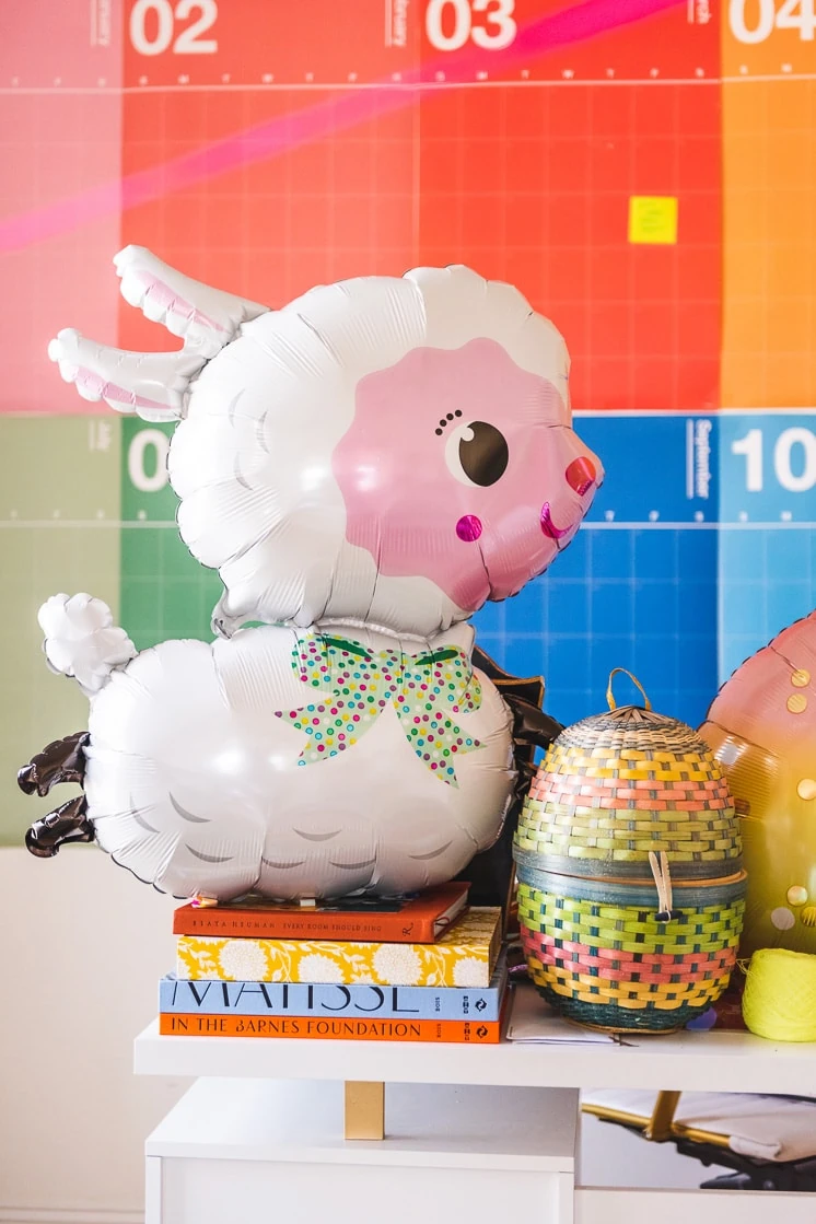 A big Easter lamb balloon rests atop a stack of colorful books in a colorful interior space. 
