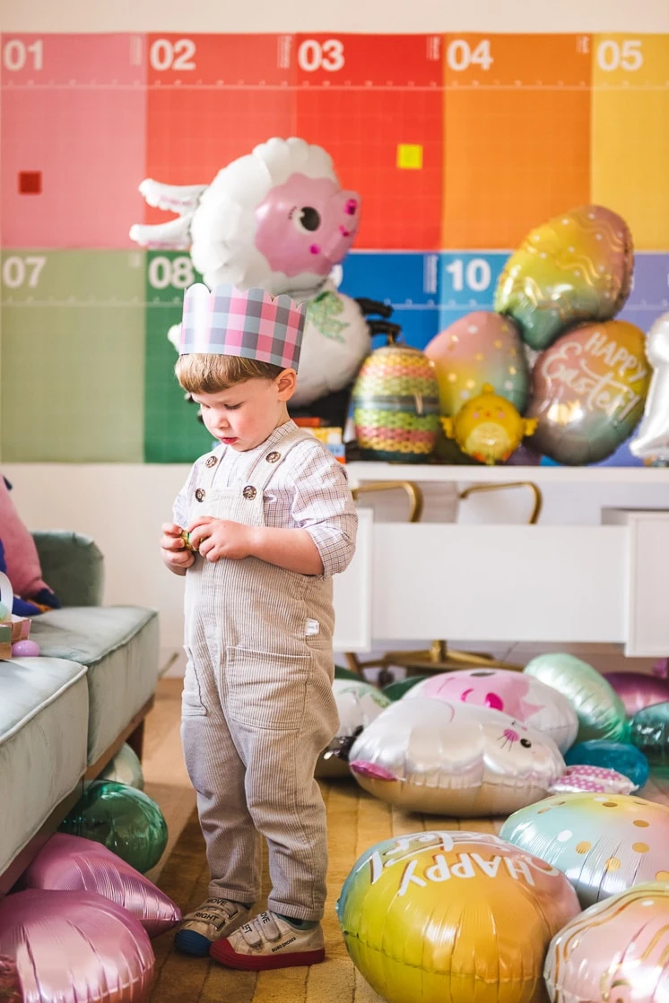A little boy in a pink and blue Easter crown stands in a colorful room filled with Easter-themed balloons.