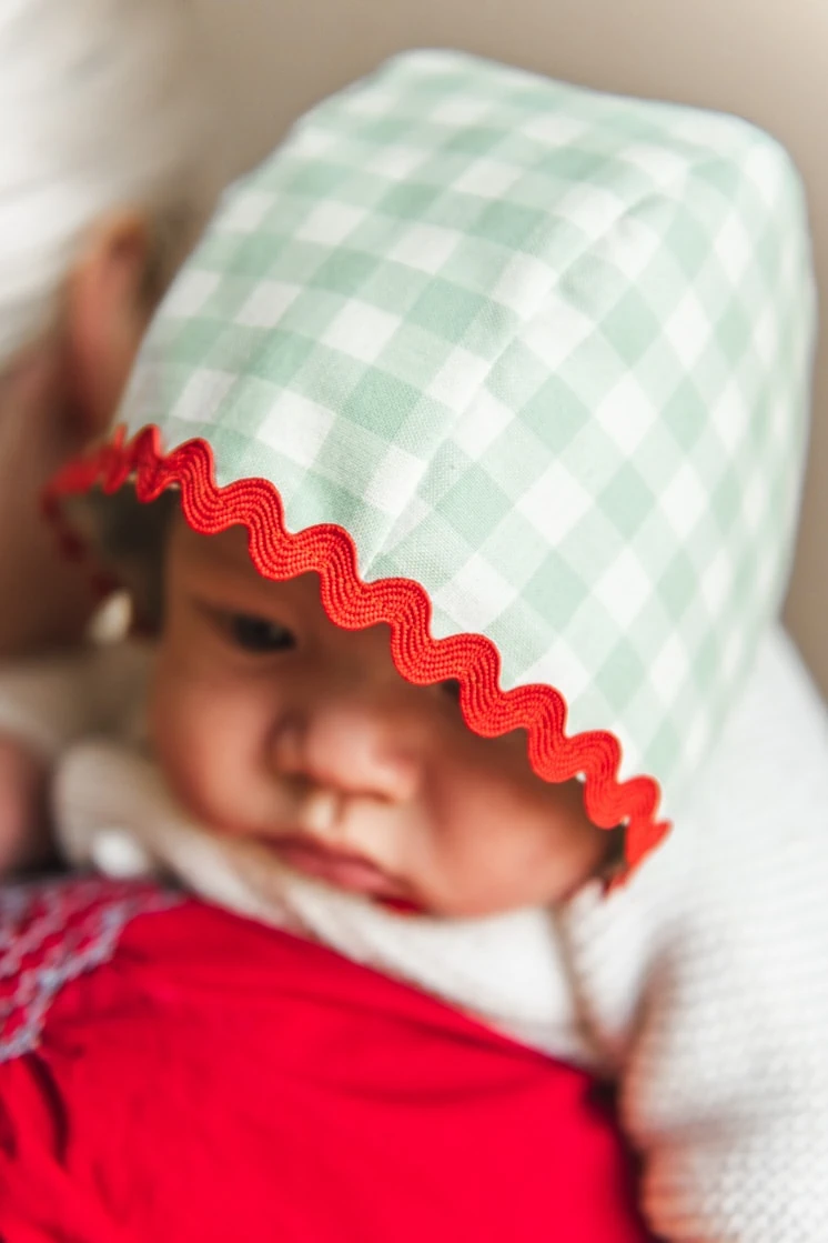Baby wearing green and red bonnet looks over woman's shoulder