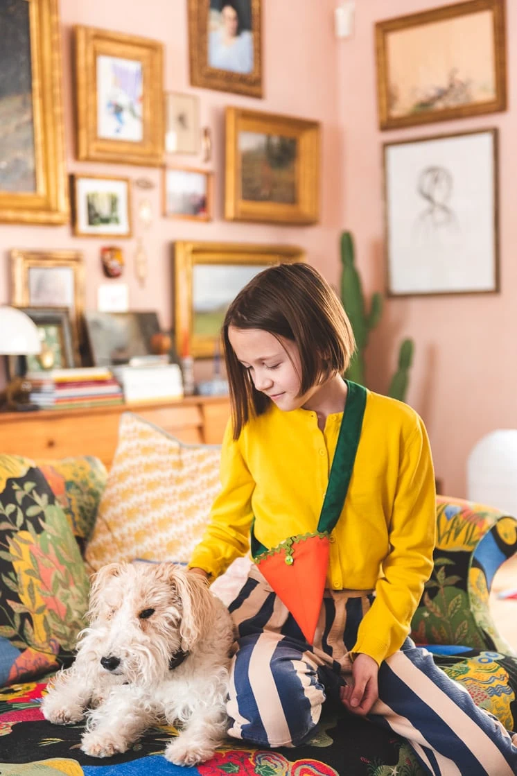 A girl in a yellow shirt pets a dog while wearing a fabric carrot bag over her shoulder.