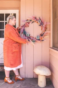 Brittany hangs up a pastel floral Easter wreath on a pink house. She's wearing a persimmon coat, a white dress, and clogs, and a cute wooden mushroom is on the porch.