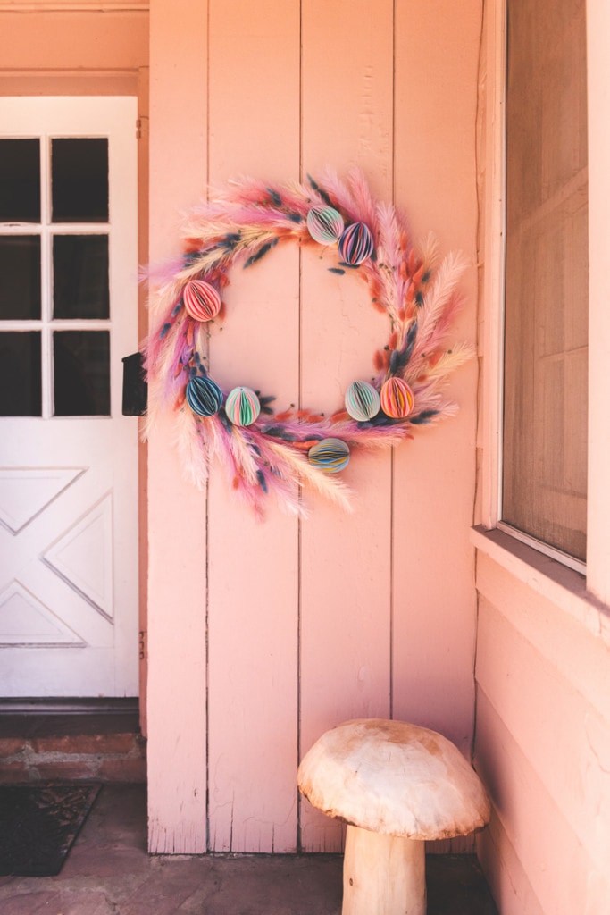 A floral Easter wreath hanging on a pink wall next to a white door. A wooden mushroom is also on the porch.