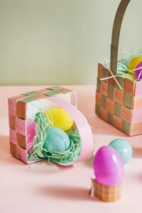 Paper grass and plastic eggs spill out of a paper Easter basket against a pink and green background