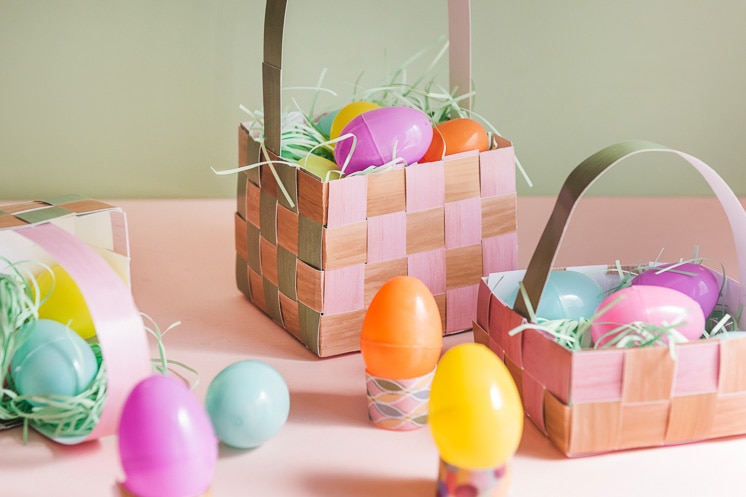 Paper Easter baskets filled with paper grass and plastic eggs against a pink and green background