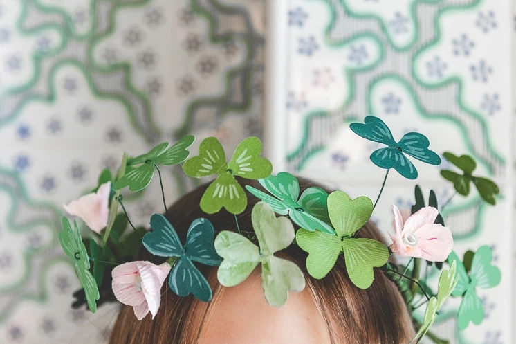 A paper shamrock and flower crown on a brunette girl's head against green and white wallpaper
