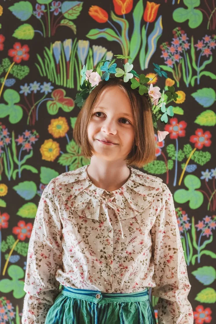 A little girl with brown hair wears a paper shamrock crown and a cream colored floral blouse. The background is bold colored wallpaper.