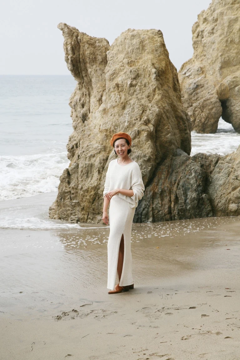 Jeanee stands on a beach wearing a white top and a white skirt. She's looking at the camera and smiling.