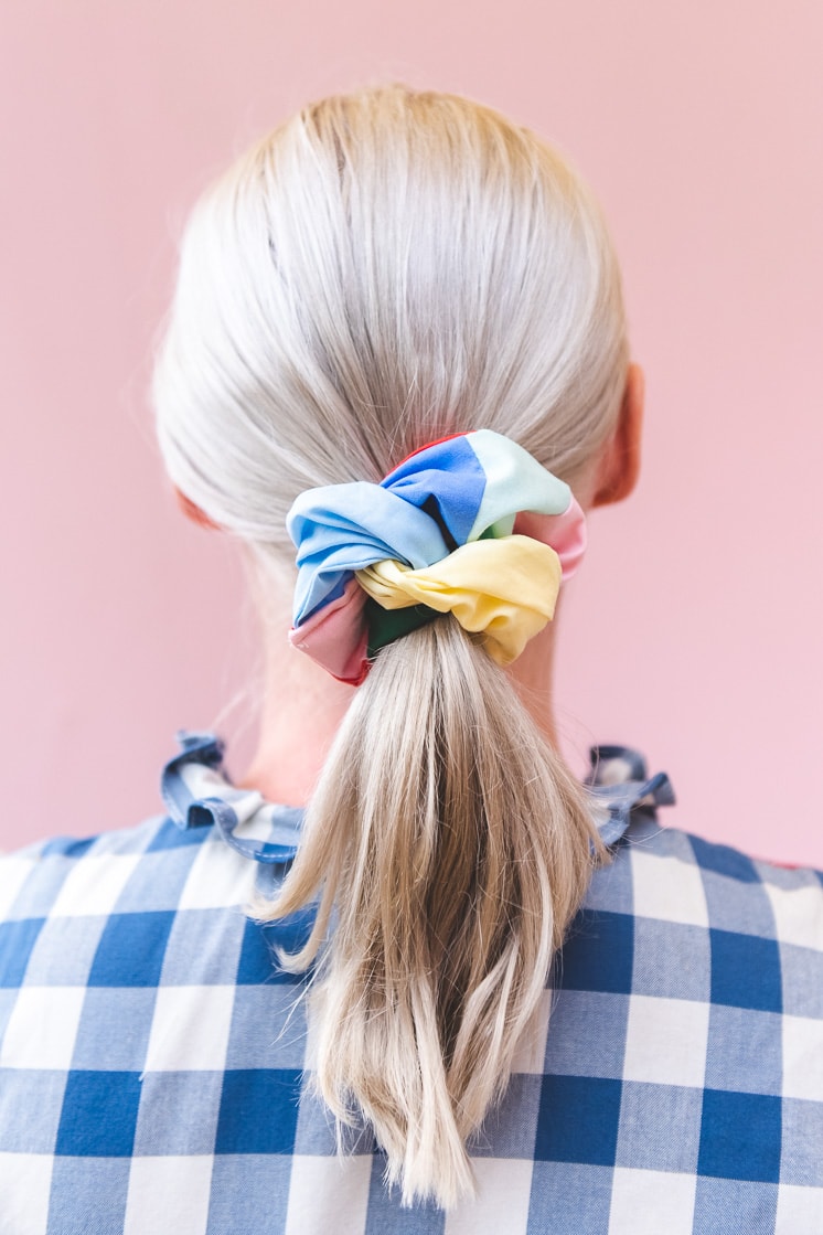 Brittany is wearing a blue and white gingham shirt and a multicolored patchwork scrunchie in her blonde ponytail. She's standing in front of a blush pink wall.