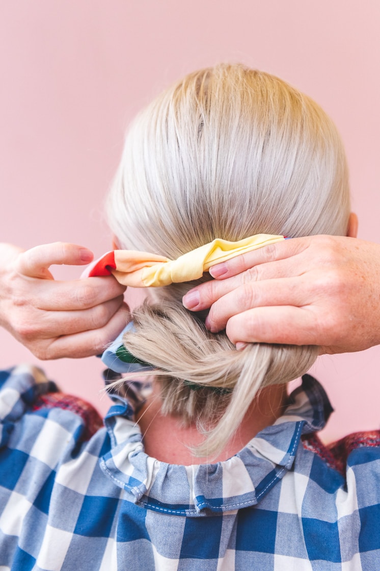 Brittany is wearing a blue and white gingham shirt and a multicolored patchwork scrunchie in her blonde ponytail. She's standing in front of a blush pink wall.