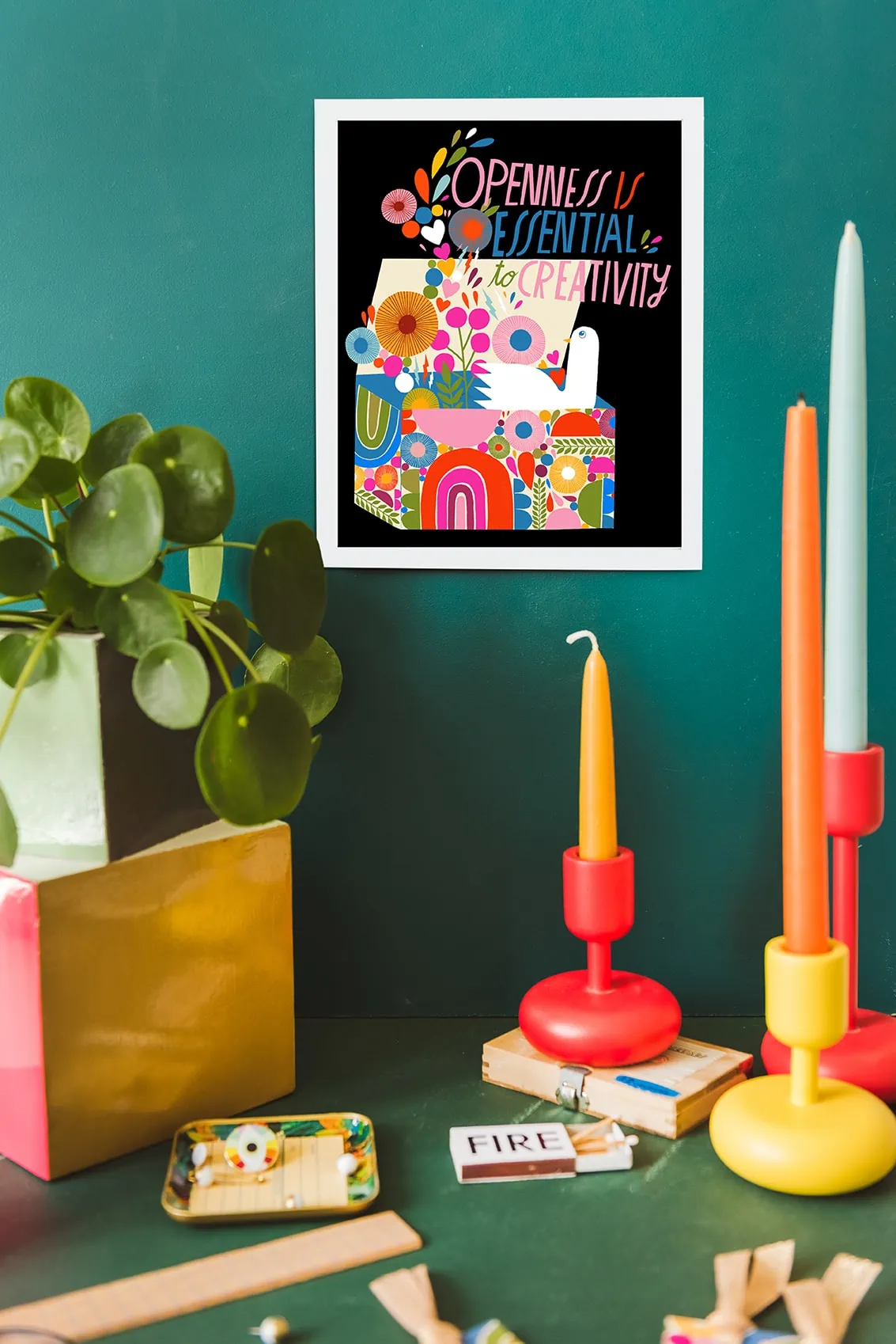 Colorful art print with the words "openness is essential to creativity" among brightly-colored candlesticks and plants in a green space. 