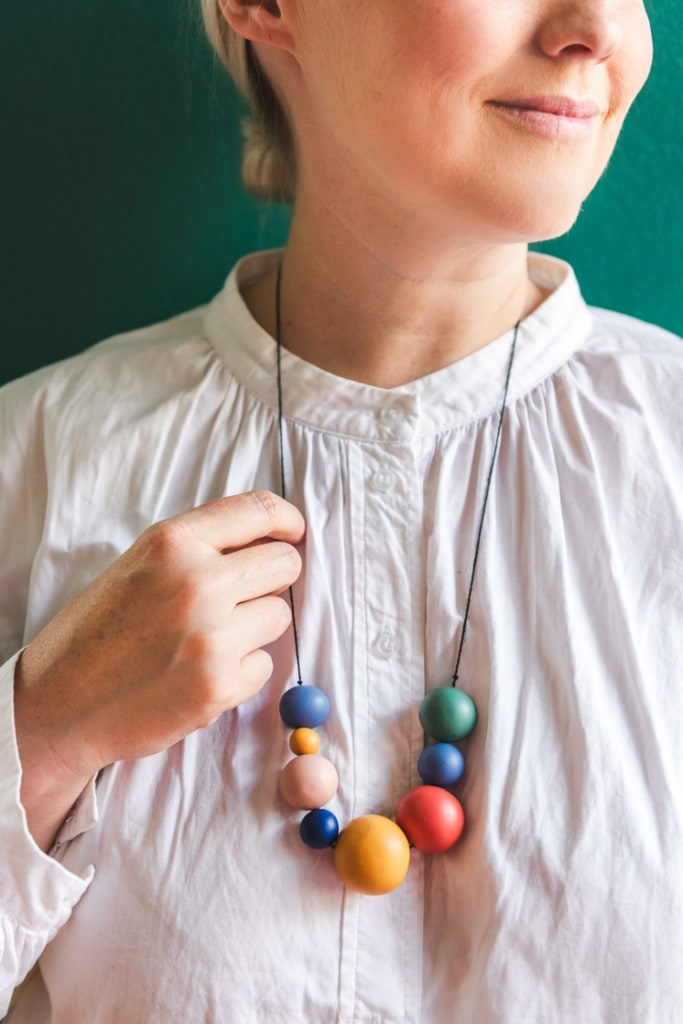 Brittany is wearing a white blouse and a rainbow colored clay necklace. She's standing against a green wall.