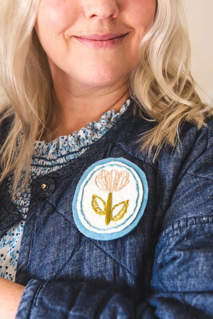 Brittany is smiling at the camera wearing a dress with a ruffled collar and a chambray quilted jacket. She's sporting her embroidered floral brooch.