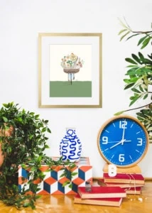 Art print of a person riding a bike with a basket full of flowers on a white wall with plants, a clock, and vases around.