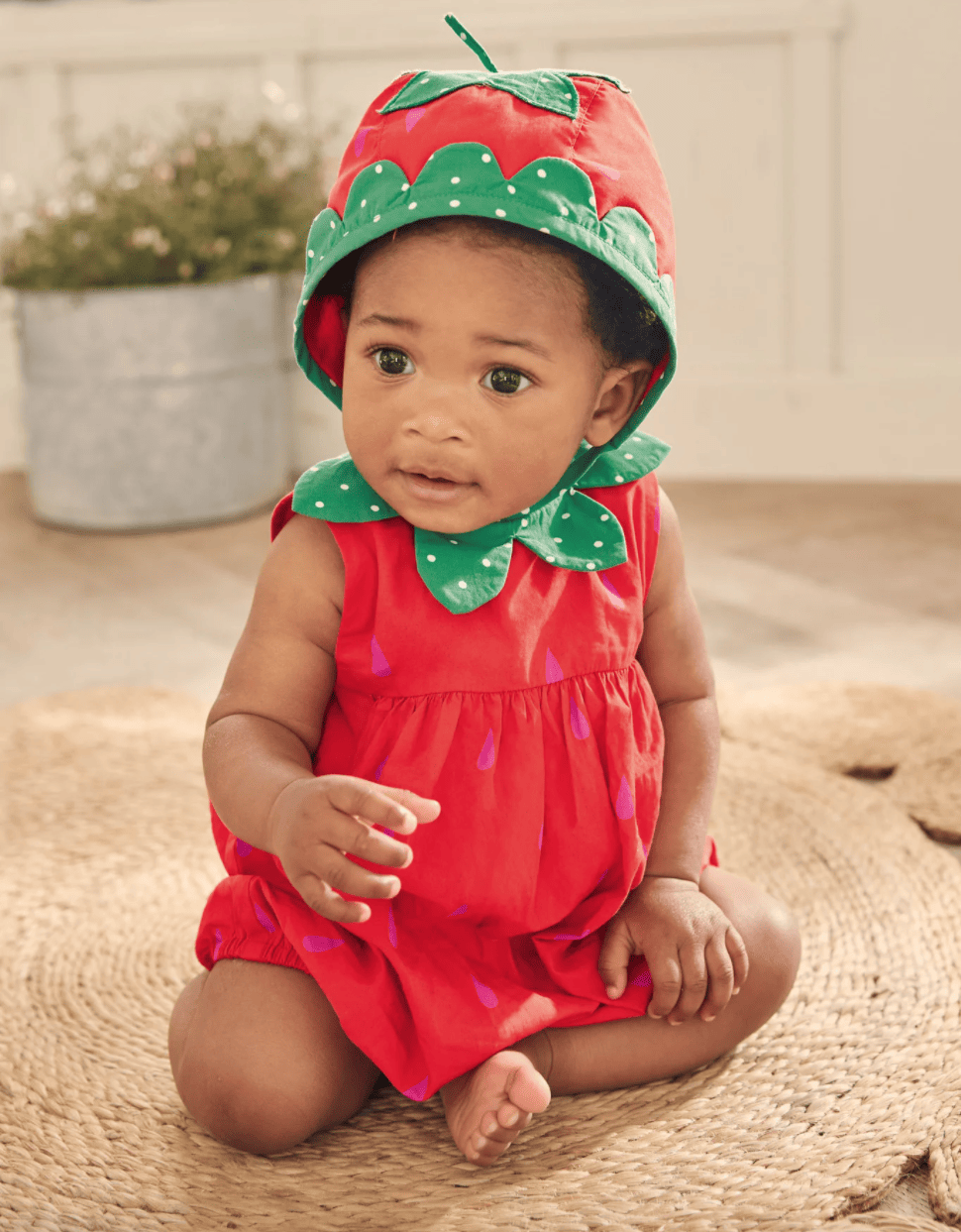 Baby wearing a strawberry bubble romper and a matching hat while sitting on a rug.