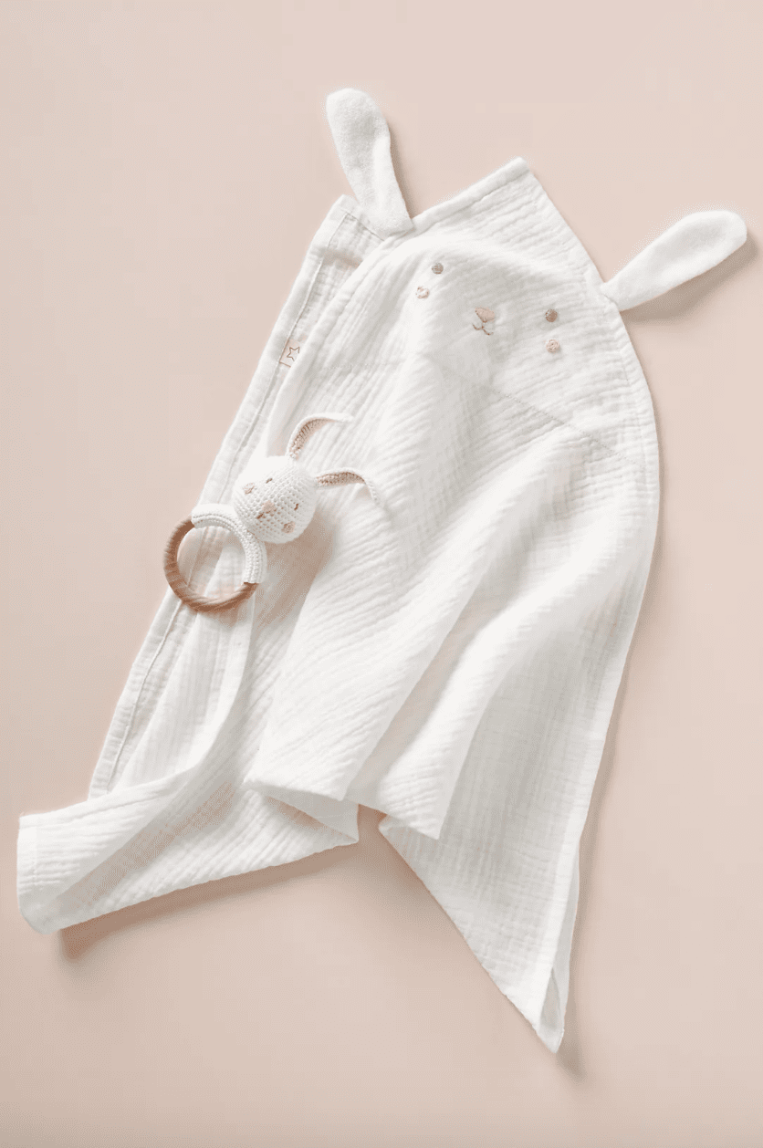 A white bunny swaddle and rattle set on a beige background. 