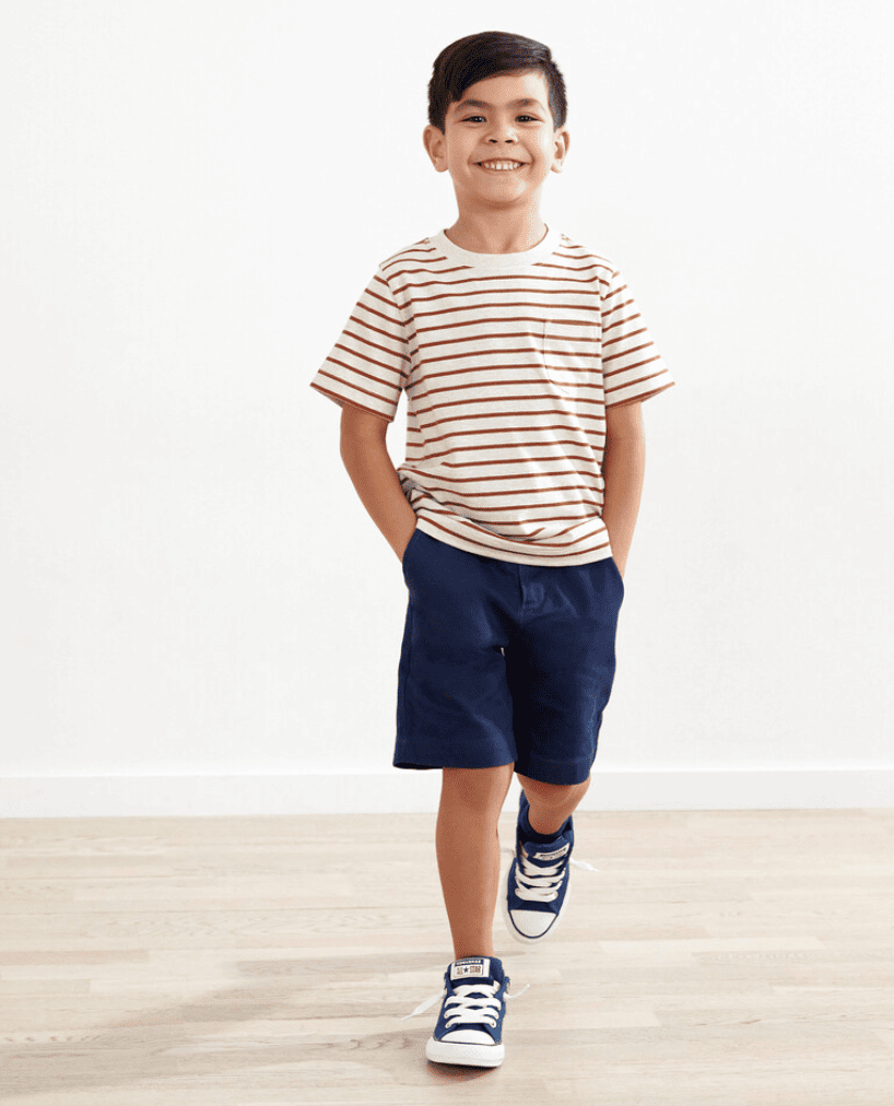 Boy wearing a cream and rust colored striped shirt and navy shorts smiles and walks toward camera