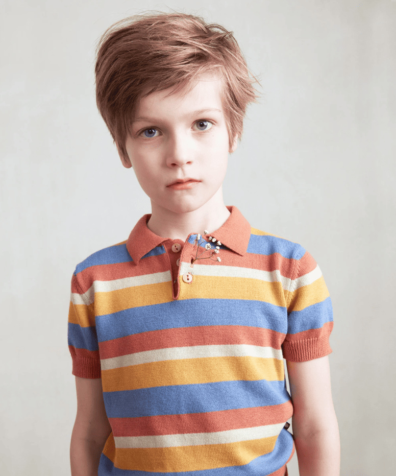 Boy looks into the camera wearing a knit striped polo in orange, cream, yellow, and cornflower blue. He has a small sprig of baby's breath tucked into his buttonhole. 