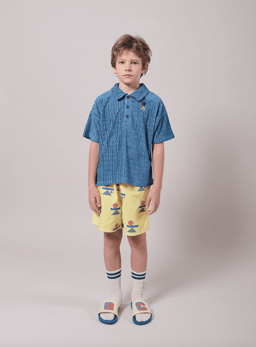 A boy in a blue polo shirt and yellow shorts with balancing shapes looks into the camera.