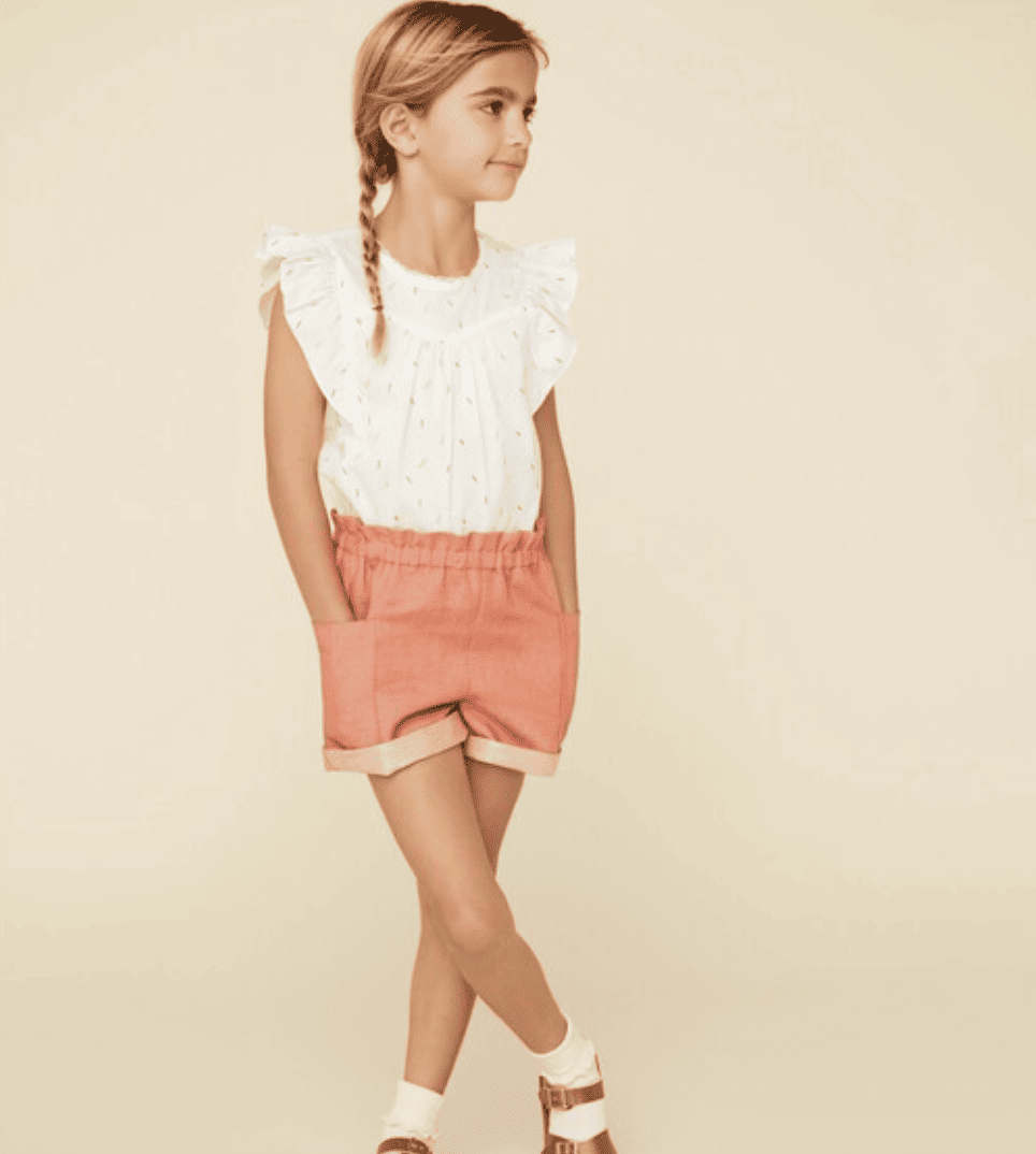 A girl wears a white ruffle sleeve blouse with multicolored sprinkles on it and salmon shorts