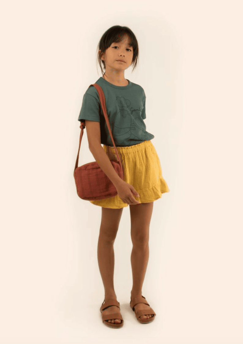 A girl wears a forest green shirt, mustard yellow frilled shorts, and sandals. 
