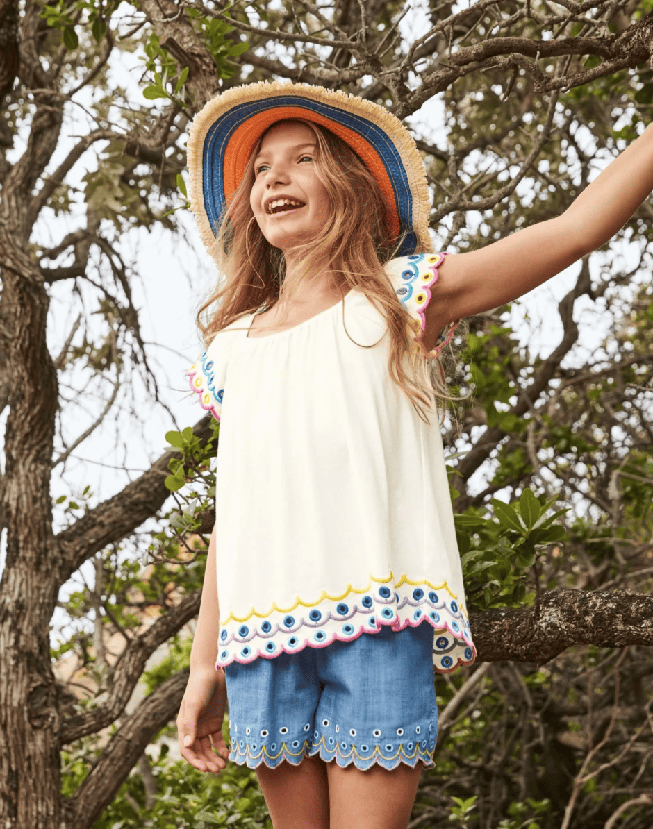 A girl wears a floppy sun hat, an embroidered blouse and chambray scalloped shorts