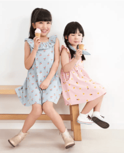 Two girls eat ice cream on a bench. The one on the left is wearing a blue ruffle neck dress with cherries printed on it.