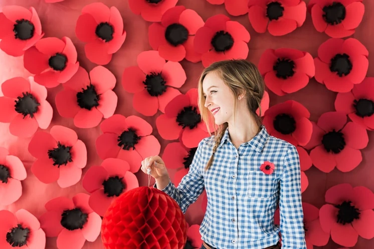 A woman holds a paper ornament in front of a wall of paper poppies