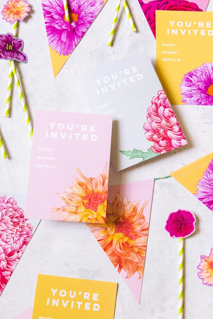 Floral party invitations with different flowers on pink, grey, and yellow backgrounds sit amongst floral bunting and cake toppers.