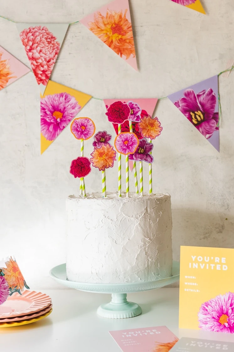 Sherbet-colored floral bunting hangs in the background. A white frosted cake with floral cake toppers sits on a mint green cake stand. Invitations and a floral crown peek in at the corners. 