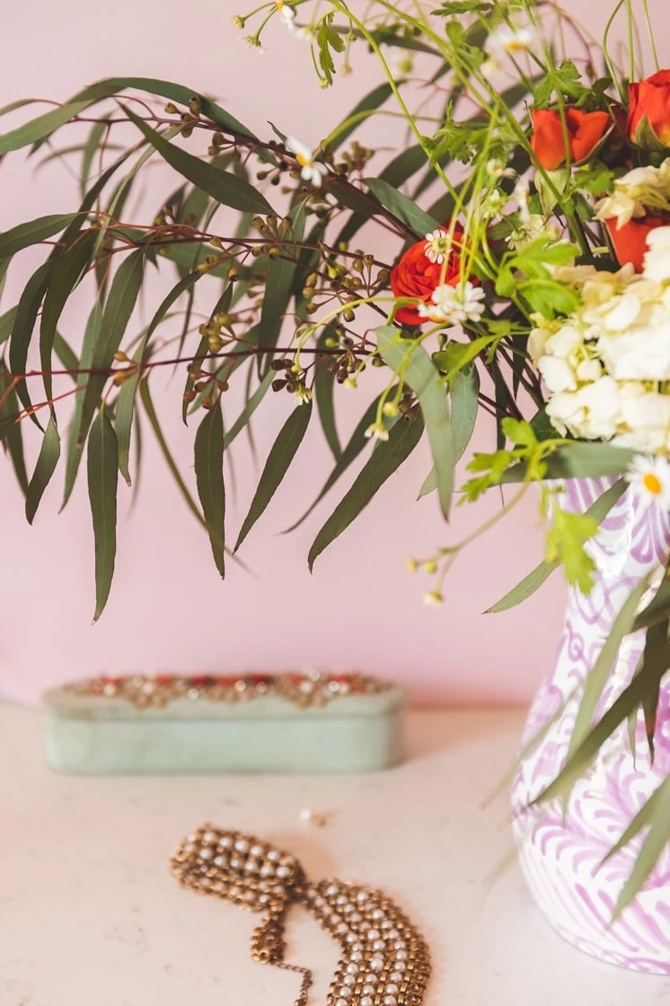 Detail shot of eucalyptus leaves, chamomile, roses, and hydrangeas against a pink wall with a mint green jewelry box in the background.