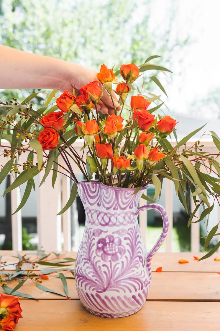 A person arranges bright orange spray roses in a purple and white vase full of eucalyptus branches.